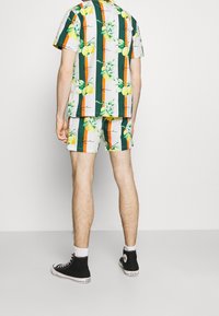 Conjunto de camisa de manga corta y pantalones cortos en tela ligera, con un colorido estampado de limones y hojas, acompañado de rayas naranjas y verdes. Zapatillas negras de caña alta.