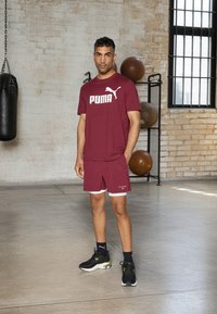 T-shirt de sport bordeaux avec logo "PUMA" blanc, short bordeaux assorti avec des accents blancs, baskets noires, et un cadre intérieur texturé.