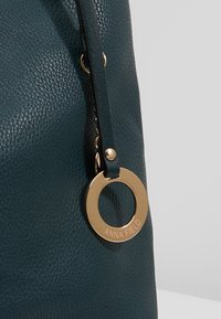 Týrkysově modrá kabělka z texturovanékůže se zlatým kruhovým doplňkem s názvem značky. Obsahuje černý popruh a kovové detaily.