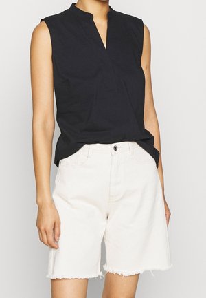 Kobieta nosząca czarny top bez rękawów z dekoltem w serek oraz wysokiej talii białe szorty jeansowe z postrzępionymi brzegami.