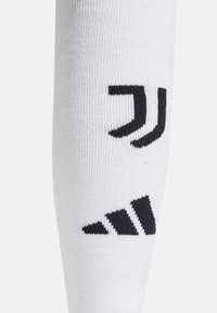 Calcetín deportivo blanco con gráficos negros. Incluye un emblema del club y un patrón de tres franjas en la zona del tobillo, hecho de un material elástico.