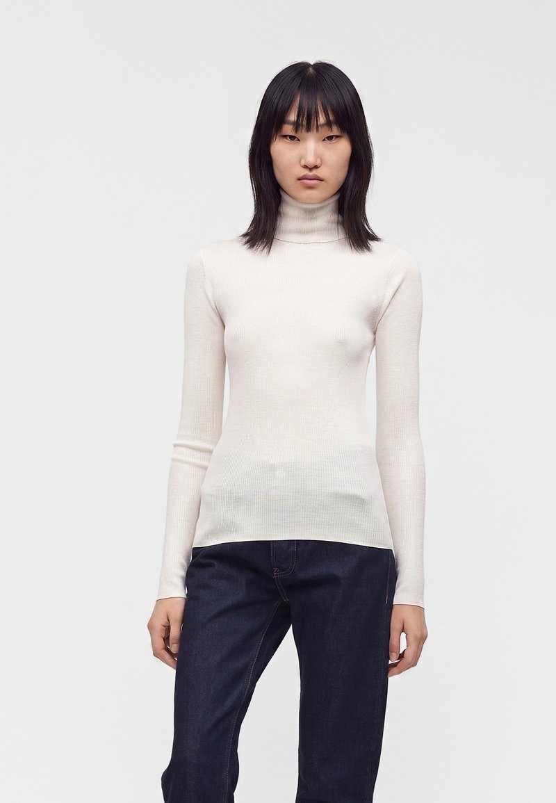 Jeune femme aux cheveux noirs raides, portant un pull à col roulé blanc et un jean bleu foncé, debout devant un fond clair uni.