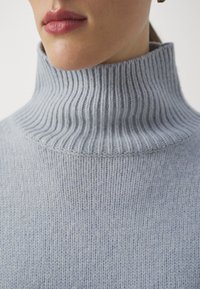 Světlé šedé žebrované rolákové svetr, který má vysoký límec a strukturovaný pletený vzor. Detailní pohled zdůrazňuje jemnost látky.