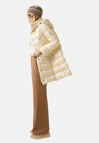 Kremowa kurtka puchowa z kapturem i zamkiem błyskawicznym, z pikowanym wzorem. Połączona z brązowymi spodniami o szerokich nogawkach i beżowym obuwiem.