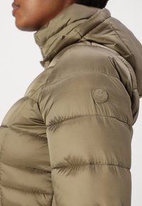 Khaki värvi puhuliikuv jakk, millel on kapuuts, horisontaalne õmblus ja ümmargune logo vasakul õlal, samuti sile ja läikiv tekstuur.