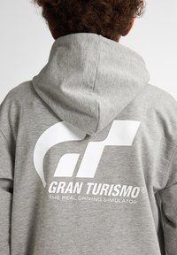 Felpa grigia realizzata in morbido materiale con un grande logo bianco e testo sul retro: "GRAN TURISMO IL VERO SIMULATORE DI GUIDA."