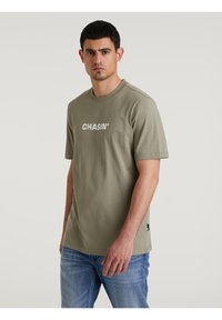 Olivezöld pamut póló rövid ujjakkal, kerek nyakkivágással és fehér "CHASIN'" logóval a mellkasán, kék farmerral párosítva.