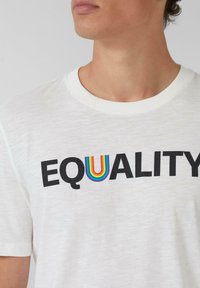 Weißes T-Shirt mit kurzen Ärmeln, auf dem das Wort "EQUALITY" in fetten schwarzen Buchstaben und einem regenbogenfarbenen "U"-Akzent in der Mitte zu sehen ist.