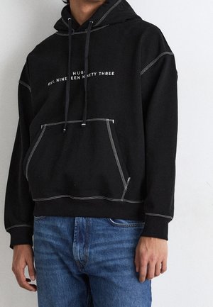 Person iført sort hættetrøje med hvid tekst og kontrastsyninger, kombineret med blå jeans, stående mod en lys baggrund.