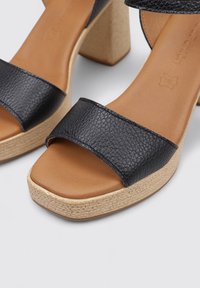 Sandalias de tacón cuadrado de cuero negro con correas anchas, plantilla de color marrón claro y plataforma tejida texturizada para agarre y estabilidad.