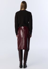 Un pull noir en peluche associé à une jupe en cuir bordeaux taille haute, avec une texture lisse et une fente à l'arrière, complété par des bottes marron jusqu'aux genoux.