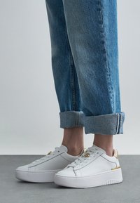 Jambes en jean bleu retroussé portant des baskets à plateforme blanches avec des accents dorés, debout sur un sol gris contre un fond clair uni.