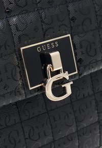 Musta patenttivahan käsilaukku, jossa on teksturoitu pinta, kohokuvioitu logokuvio, suorakulmainen kultasävyinen solki ja kultainen "G"-koriste.