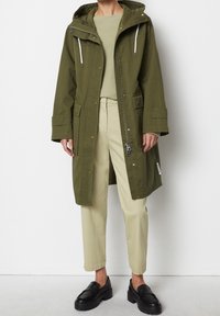 Olivgrön vattentät parka med huva, dragkedja och tryckknappar, två framfickor och justerbara dragsko. Kombinerad med krämfärgade byxor och svarta skor.