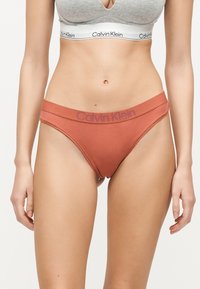 Femme portant un soutien-gorge de sport gris Calvin Klein et un slip bikini couleur rouille Calvin Klein sur un fond uni.