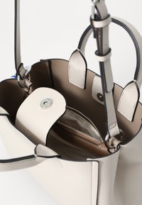 Sac à main en cuir beige clair avec une texture lisse, comportant une poche zippée à l'intérieur et des bords contrastants sombres. Deux poignées supérieures et une sangle réglable.