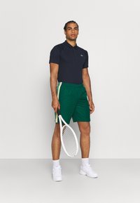 Camisa de polo azul marino, pantalones cortos verdes con lados a rayas, zapatillas de tenis blancas y sosteniendo una raqueta de tenis. Fondo liso.