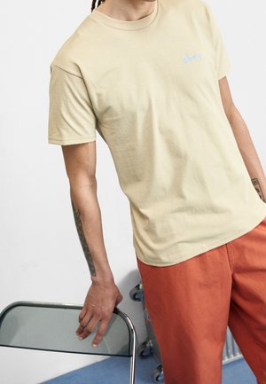 Beiges Baumwoll-T-Shirt mit kurzen Ärmeln und rundem Halsausschnitt, mit einem hellblauen "Obey"-Logo auf der Brust, kombiniert mit orangen Hosen.