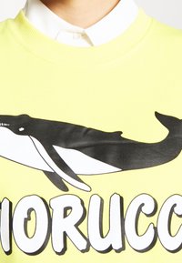 Sudadera amarilla brillante con un gráfico de ballena negra, escote redondo y puños de canalé. El texto debajo dice "Miorucco" en negrita y fuente negra.