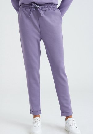 Lila Sweatpants mit einem Kordelzugbund, schmal zulaufenden Beinen und gerippten Knöcheln, aus einem weichen Stoff gefertigt. Weiße Sneakers runden den Look ab.