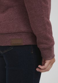 Maroon dukserica od teksturiranog materijala, s rebrastim rukavima i smeškim kožnim logom na donjem rubu, u kombinaciji s tamnoplavim farmerkama.
