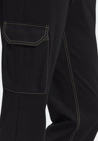 Black pants with a side pocket, featuring yellow stitching along the seams and a textured fabric. Pocket has a rectangular shape and flap closure.