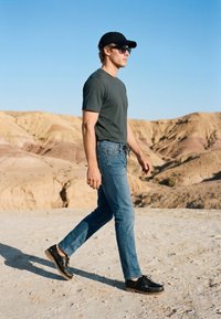 Homme portant une casquette noire, des lunettes de soleil, une chemise sombre et un jean marchant sur un terrain rocheux avec des collines désertiques sous un ciel bleu clair.