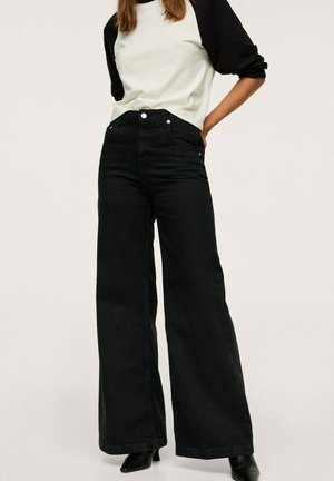 Femme portant un jean noir à jambes larges, un t-shirt raglan noir et blanc, et des bottes noires à bout pointu, debout, une main sur la hanche.