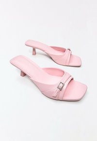Paire de mules roses à talons ouverts avec bouts carrés et petites boucles sur fond blanc.