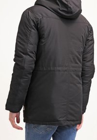 Svart puffjacka med huva, med kviltat stygn och en slät yta. Bärs över blå jeans, med en etikett vid fållen.