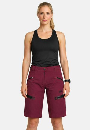 Frau steht und trägt ein schwarzes ärmelloses Oberteil sowie knieumspielende, kastanienbraune Shorts mit Reißverschlusstaschen und eine Armbanduhr am linken Handgelenk.