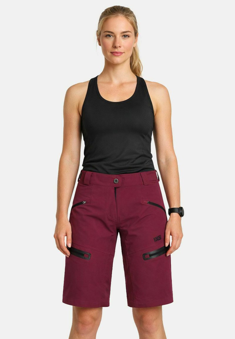 Femme debout portant un haut noir sans manches et un short bordeaux à hauteur des genoux avec poches à fermeture éclair, ainsi qu'une montre-bracelet au poignet gauche.