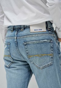 Jeans de mezclilla azul claro con un acabado desgastado, con costuras amarillas, dos bolsillos traseros y una etiqueta blanca con "SALSA JEANS."