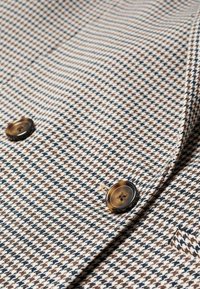 Tela de patrón pata de gallo en tonos marrón claro y negro, con doble botonadura, dos botones redondos de color marrón oscuro y textura suave.