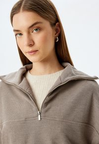 Vaalean beige ribattu puolivetoketjupaita, joka on valmistettu pehmeästä kankaasta ja jossa on käännettävä kaulus sekä hopeiset yksityiskohdat.
