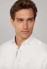 Camisa de lino blanca con un collar estructurado, que presenta acentos a rayas azules y blancas a lo largo de la tapeta de botones y una textura suave.