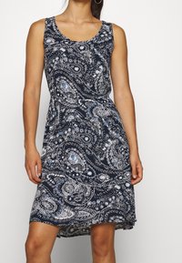 Femme portant une robe sans manches bleu marine avec des motifs paisley et floraux blancs et bleu clair, se tenant devant un fond uni.