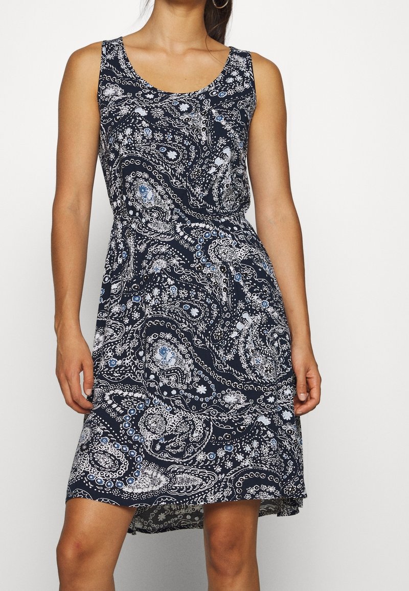 Femme portant une robe sans manches bleu marine avec des motifs paisley et floraux blancs et bleu clair, se tenant devant un fond uni.