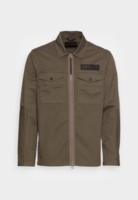 Olivengrøn jakke med lynlås, to brystlommer og et påsyet mærke. Fremstillet af holdbart stof med en glat tekstur.