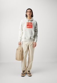 Hoodie blanc avec graphique rouge, pantalon beige et baskets marron/crème. Tenant un sac à dos beige avec poches zippées et lignes épurées.