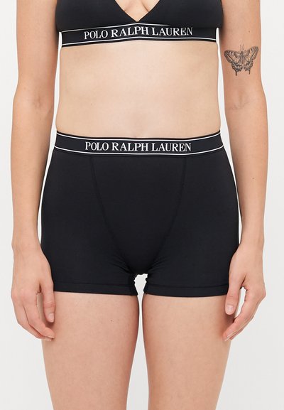 Vrouwelijk bovenlichaam in een zwarte sportbeha van Polo Ralph Lauren met bijpassende boxershort, vlindertattoo zichtbaar op de linkerarm.