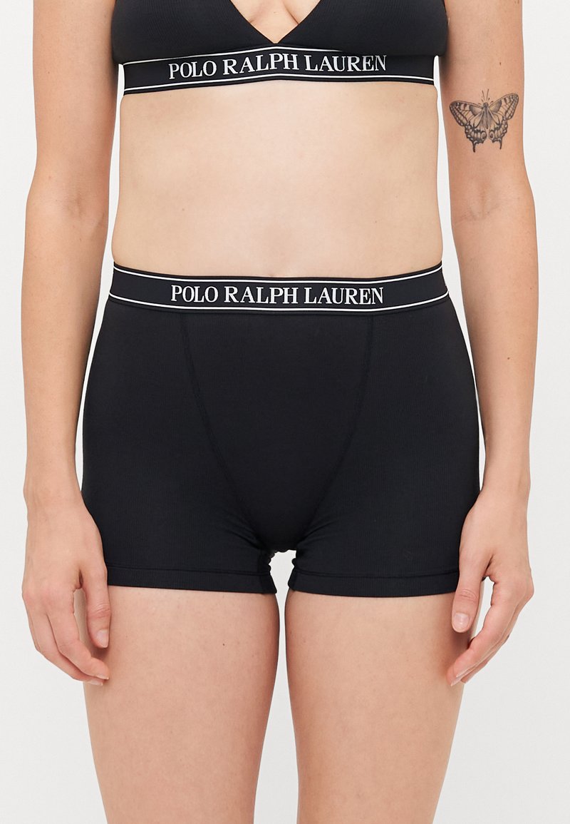 Kvinnekropp iført svart Polo Ralph Lauren-merket sportbh og matchende boxershorts, sommerfugltatovering synlig på venstre arm.