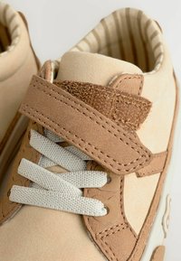 Baskets en daim beige avec des accents marron, des lacets blancs et une sangle Velcro. L'intérieur est doublé d'un tissu rayé.