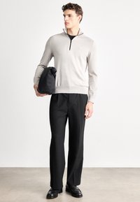 Helehall zipiga kampsun musta kraega, mustade volditud pükste ja mustade kingadega, käes tume kott.