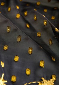 Juodas audinys su tekstūruota paviršiumi, papuoštas auksinėmis siuvinėtomis motyvais, kurie kartojasi. Lygus, permatomas medžiaga su subtiliu blizgesiu.