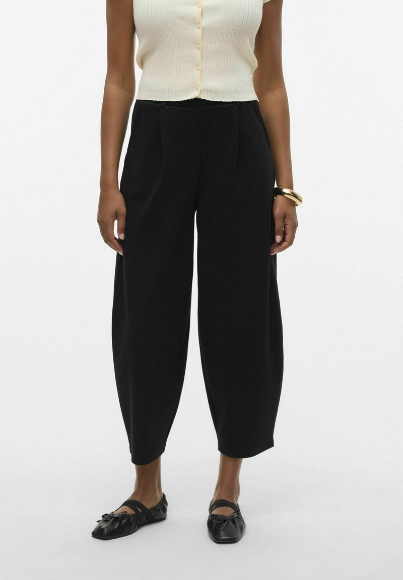 Pantalon noir taille haute, à jambes larges avec plis et texture lisse, associé à des chaussures plates noires et un top crème à côtes, boutonné.