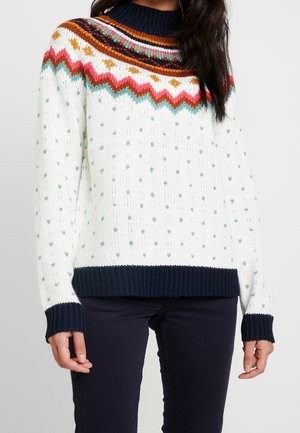 Pull en maille blanc avec des pois bleu sarcelle, motif zigzag multicolore sur l'empiècement, poignets, ourlet et col en côtes bleu marine, porté par une personne en pantalon foncé.
