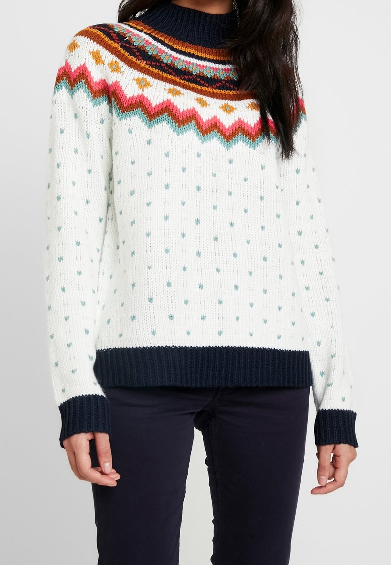 Pull en maille blanc avec des pois bleu sarcelle, motif zigzag multicolore sur l'empiècement, poignets, ourlet et col en côtes bleu marine, porté par une personne en pantalon foncé.