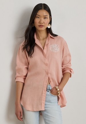 Kobieta o długich ciemnych włosach, ubrana w luźną różową koszulę zapinaną na guziki z białym monogramem oraz jasnoniebieskie dżinsy, stojąca na jednolitym tle.