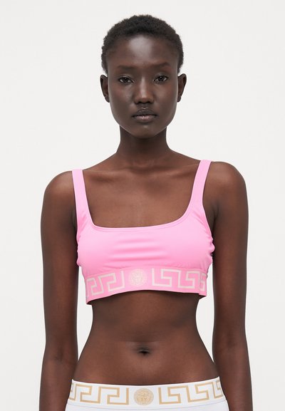 Roze sportbeha met vierkante halslijn, dikke bandjes en een gouden Grieks sleutelpatroon langs de onderkant. Gemaakt van een gladde, rekbare stof.